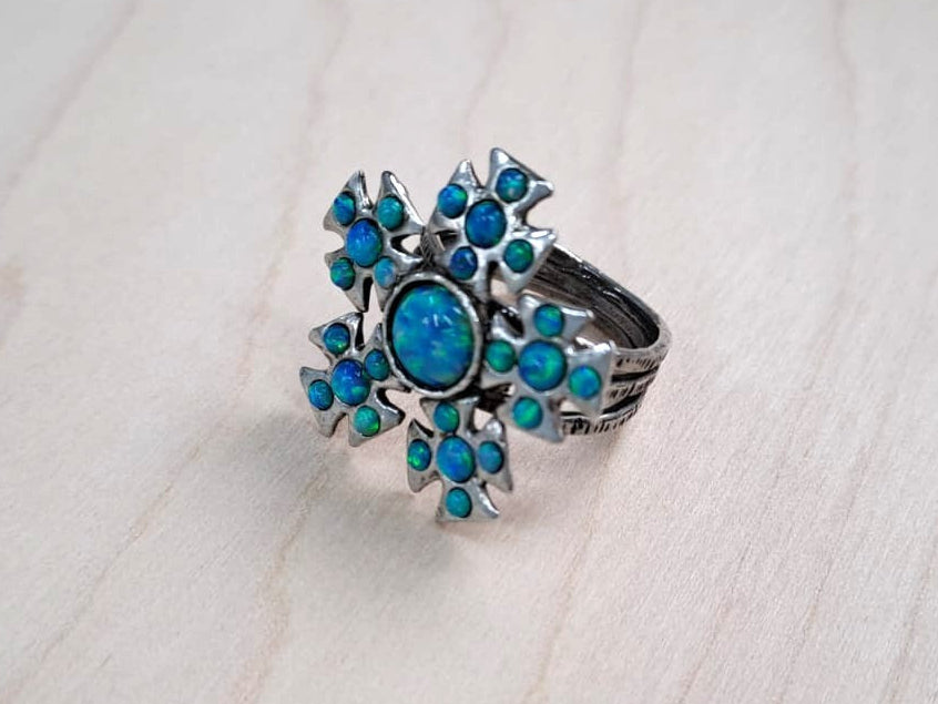 A silver-toned ring with a floral cluster setting featuring bright blue stones, including a central round blue stone, resting on a light-colored wood surface.