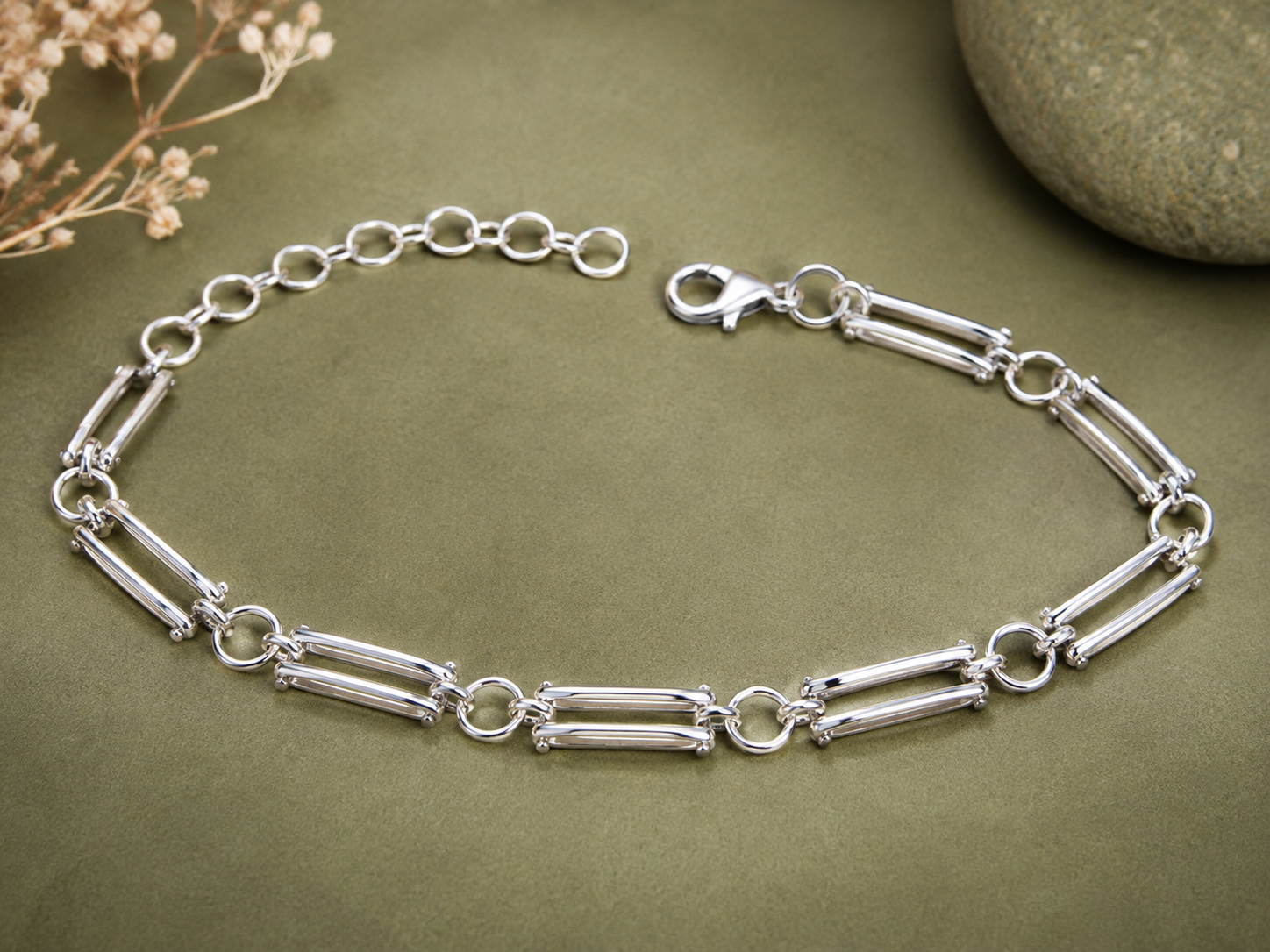 A silver-toned chain bracelet with alternating cylindrical links and small circular links, displayed on an olive green textured surface with dried flowers and a stone in the corners.